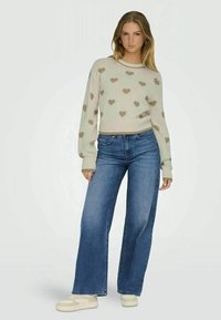 Sweater de malha bege com padrões de corações, combinado com jeans de pernas largas azuis. Modelo usa ténis de cor clara. Texturas suaves por toda a peça.