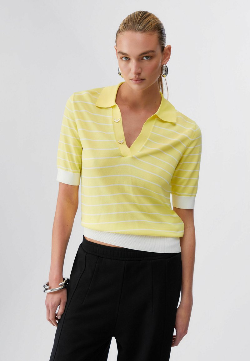 Femme aux cheveux blonds portant un polo à manches courtes rayé jaune et blanc et un pantalon noir, accessoirisée avec des boucles d'oreilles et des bracelets en argent.
