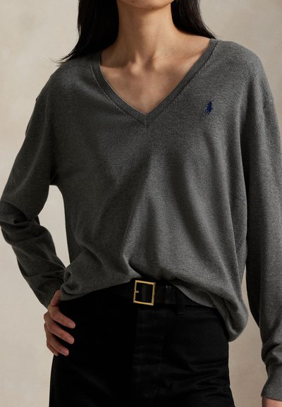 Moteris vilki pilką V formos kaklo megztinį ilgomis rankovėmis, juodas kelnes ir juodą diržą su auksine sagtimi, ranka laikosi už klubo.