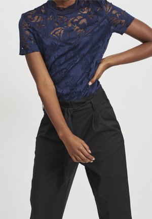 Vrouw draagt een marineblauw kanten top met korte mouwen en een zwarte broek met hoge taille, poseert met één hand op haar heup en de andere ontspannen langs haar lichaam.