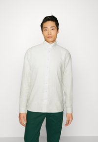 Camisa de lino clara de botones con cuello, mangas largas y dobladillo recto, combinada con pantalones de color verde oscuro.