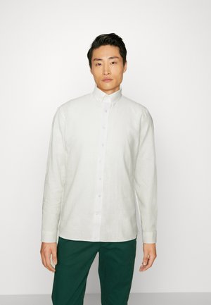 Light-colored linen button-up shirt with a collar, long sleeves, and a straight hem, paired with dark green trousers.