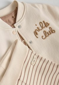 Gilet beige avec col côtelé, boutons-pression, et "milk club" brodé en marron. Présente des manches rayées et une texture douce.