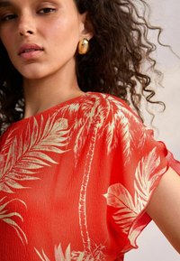 Red sheer top featuring a palm tree pattern. The fabric has a crinkled texture, with loose, short sleeves and a wide neckline. Gold earrings visible.