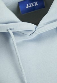 Felpa blu chiaro realizzata in tessuto morbido, con cappuccio regolabile e un'etichetta di marca visibile nella zona del collo. Texture liscia, design semplice.