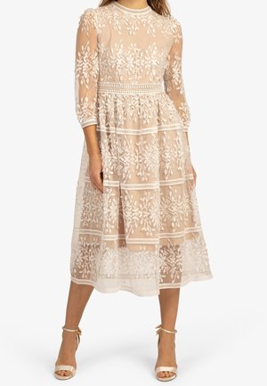 Robe beige transparente longueur midi avec broderies florales blanches, manches trois-quarts, jupe à volants, et escarpins à bride cheville assortis.