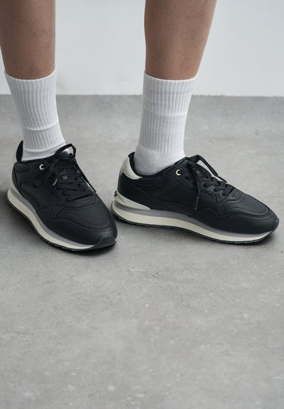 Scarpe sportive nere con tomaia in pelle texturizzata, soletta bianca e suola in gomma nera, abbinate a calzini bianchi a coste.