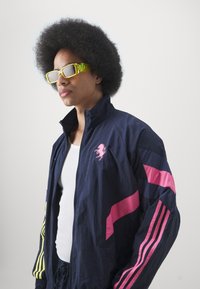 Marineblå atletisk jakke med lyserøde og gule striber, med et lyserødt broderet logo, båret over en hvid top. Suppleret med lysgule briller.