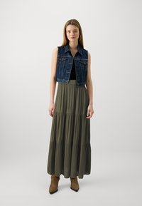 Gilet en denim avec un col sur un top noir, associé à une longue jupe tiered verte olive et des bottes chevilles marron, sur fond blanc.