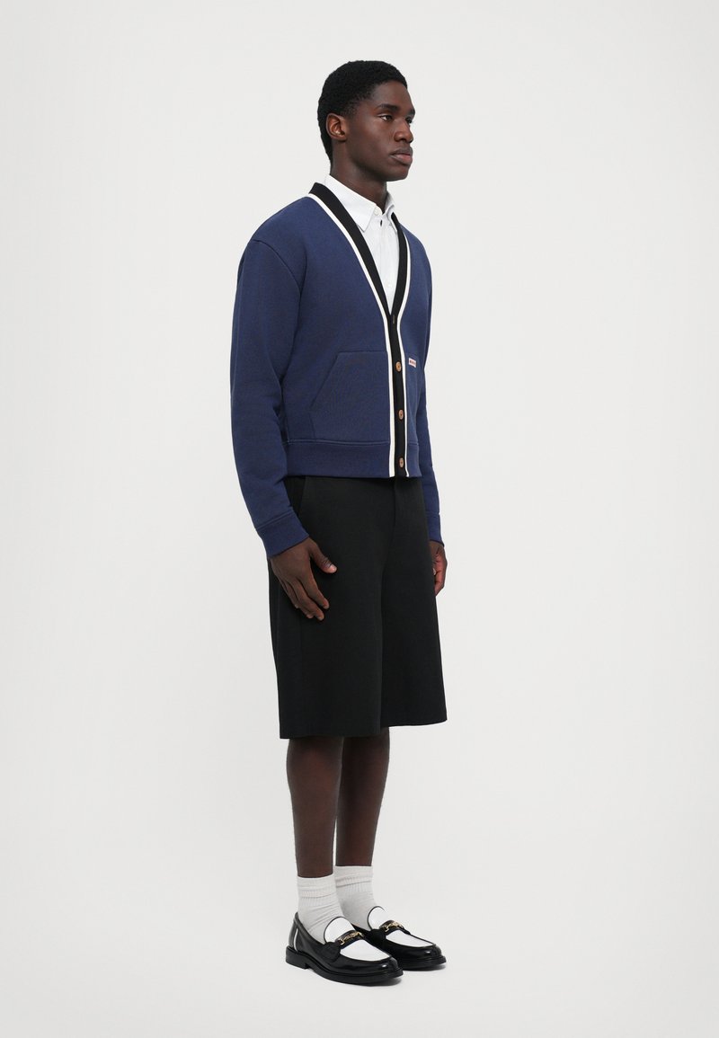 Cárdigan navy con ribete blanco, dos bolsillos delanteros y botones de madera, combinado con pantalones cortos negros y zapatos negros con acentos blancos.