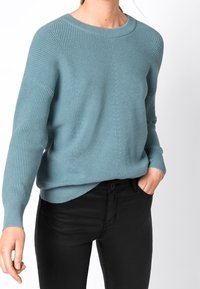 Femme portant un pull en maille côtelée bleu clair et un pantalon noir, avec une main dans la poche, sur fond blanc uni.