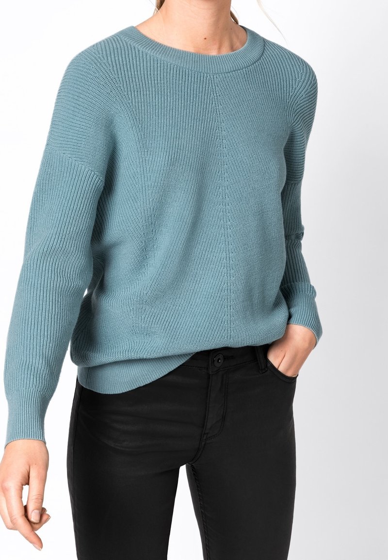 Femme portant un pull en maille côtelée bleu clair et un pantalon noir, avec une main dans la poche, sur fond blanc uni.