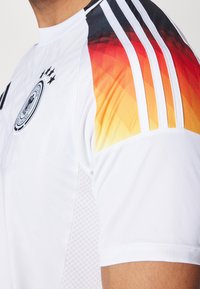 Biała koszulka sportowa z teksturowanym wzorem, z gradientowym designem w odcieniach pomarańczowego i czarnego na ramionach, trzema czarnymi paskami i emblematem.