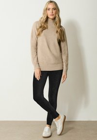 Beige neulepaita korkealla kaula-aukolla, yhdistettynä slim-fit mustiin legginsseihin ja kermaisiin lenkkareihin, joissa on ruskeat pohjat, neutraalia taustaa vasten.