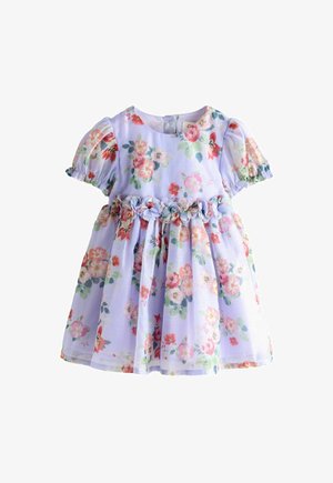 Nasmešana djevojka kovrčave kose u cvjetnoj haljini i bijelim cipelama stoji ispred štanda s cvijećem s izrezanim likovima crtanih pasa.