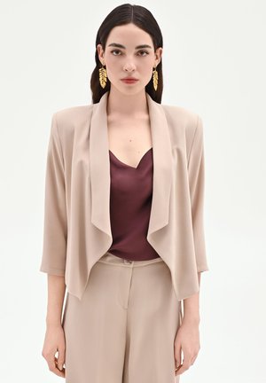 Mannequin femme aux longs cheveux foncés portant un blazer beige, un pantalon assorti, un top en satin bordeaux et des boucles d’oreilles dorées en forme de feuille sur fond blanc.