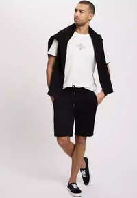 Czarne szorty z elastycznym pasem, biała koszulka z nadrukiem oraz czarny sweter zarzucony na ramionach. Całość uzupełniają czarne sneakersy.