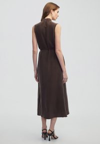 Hnědé maxi šaty bez rukávů s vysokým límcem, nařaseným pasem, hladkým materiálem a splývavým střihem, doplněné o sandály na podpatku.