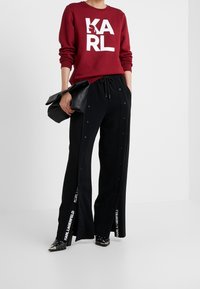 Bordeaux sweatshirt med vit "KARL" logotyp; svarta byxor med sidoknappar, dragsko i midjan och vit branding på benen. Svarta spetsiga skor.