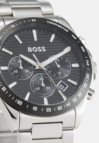 Montre ronde noire avec un cadran texturé, des accents argentés et trois sous-cadrans. Équipée d'un bracelet en acier inoxydable et d'une lunette tachymétrique.