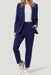 Blazer bleu marine sur mesure et pantalon assorti, en tissu lisse. Portés avec un t-shirt blanc et des baskets blanches, sans motifs.