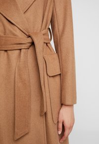 Gros plan sur un manteau en laine couleur chameau avec une ceinture nouée et une poche à rabat, porté sur le bras gauche par une personne à la main détendue.