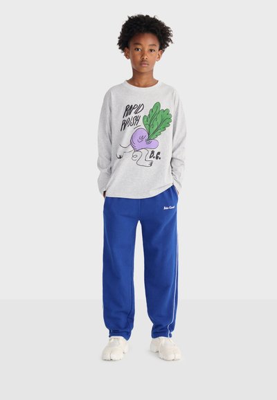 Bambino che indossa una maglia grigia a maniche lunghe con una grafica di un ravanello cartoon e pantaloni della tuta blu, in piedi davanti a uno sfondo semplice.