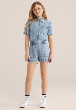 Fată tânără cu păr blond lung, purtând ochelari rotunzi, un salopetă din denim albastru deschis cu mâneci scurte, șosete în dungi și adidași albi înalți.