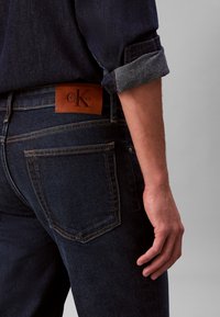 Σκούρο μπλε τζιν με διακριτή υφή. Διαθέτει καφέ δερμάτινο ταμπελάκι με λογότυπο στη μέση και κανονικές πίσω τσέπες.