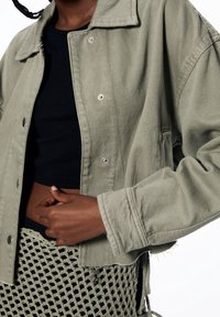 Olijfgroene kortere jas van gestructureerde stof, voorzien van een knoopsluiting aan de voorkant en een losse pasvorm. Zwart topje eronder zorgt voor contrast.