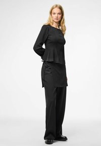 Top peplum nero con maniche lunghe e pieghe frontali, abbinato a pantaloni ampi coordinati. Tessuto liscio e texture, design semplice.