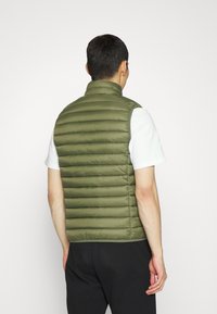 Chaleco acolchado verde oliva con cuello alto, diseño de rombos horizontal, material ligero y silueta ajustada; usado sobre una camisa blanca.