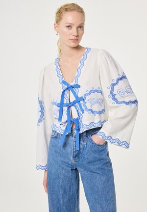 Vrouw met een witte blouse met blauwe borduursels en linten, gecombineerd met een blauwe spijkerbroek met hoge taille, staand tegen een effen witte achtergrond.