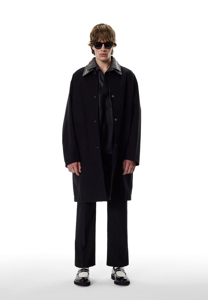 Abrigo negro de gran tamaño con cuello de cuero, llevado sobre un atuendo oscuro. Lleva gafas de sol negras y zapatos negros con detalles en blanco.