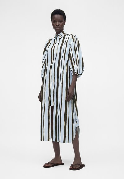 Γυναίκα που φοράει μακρύ κουμπωτό φόρεμα με κατακόρυφες καφέ και λευκές ρίγες και φουσκωτά μανίκια, ξυπόλητη σε απλό φόντο.