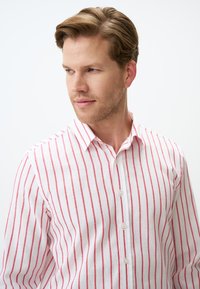 Chemise en lin blanc avec de fines rayures verticales rouges, un col pointu et une fermeture à boutons, avec une texture lisse et une coupe ajustée.