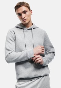 Szary hoodie z miękkiego materiału, z kieszenią kangurką, regulowanym sznurkiem i ribbami przy mankietach. Model pokazany z rękami umieszczonymi blisko klatki piersiowej.