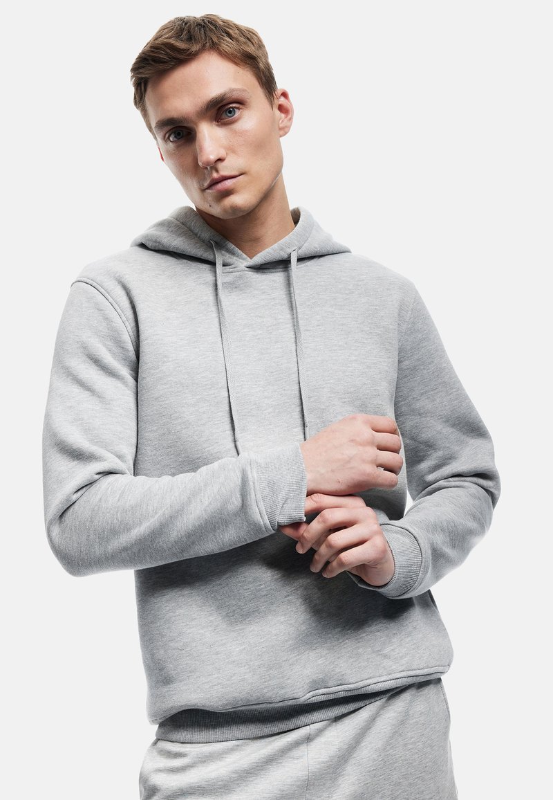 Szary hoodie z miękkiego materiału, z kieszenią kangurką, regulowanym sznurkiem i ribbami przy mankietach. Model pokazany z rękami umieszczonymi blisko klatki piersiowej.