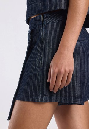 Close-up van een persoon die een donkerblauwe pinstripe rok met een zijrits draagt en een hand die tegen de dij rust.