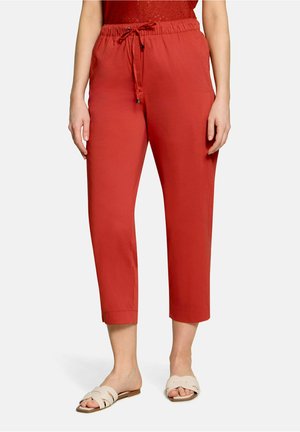 Femme portant un pantalon rouge court à cordon et des sandales beiges sans bride, debout sur un fond blanc uni.