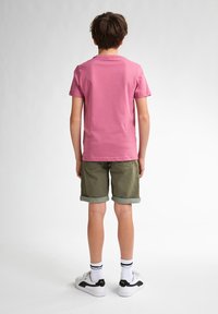 Camiseta rosa de manga corta combinada con pantalones cortos verde oliva. Zapatos blancos con detalles negros, puños enrollados y calcetines a rayas. Diseño minimalista.