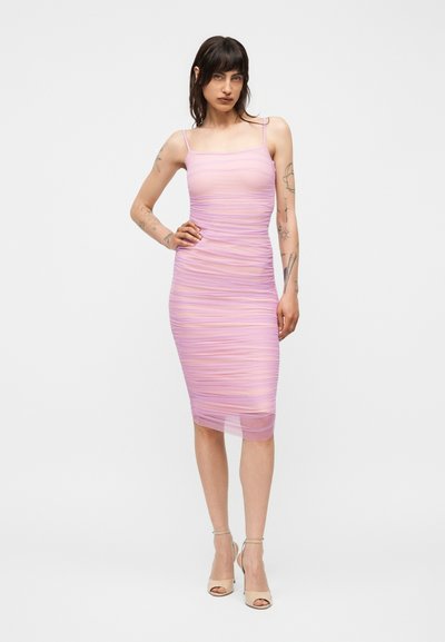 Γυναίκα με σκούρα μαλλιά και τατουάζ που φοράει εφαρμοστό, ραμμένο, αμάνικο μίντι φόρεμα σε ανοιχτό μωβ χρώμα και μπεζ ψηλοτάκουνα σανδάλια.