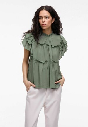 Green tiered blouse featuring ruffled accents, a high neckline, and short sleeves, made of lightweight fabric. Paired with light-colored pants.