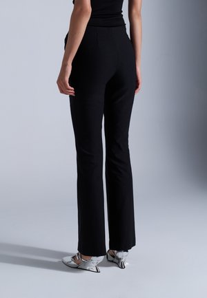 Personne portant un pantalon noir ajusté et des chaussures à talons bas métalliques argentées, debout devant un fond uni gris clair.