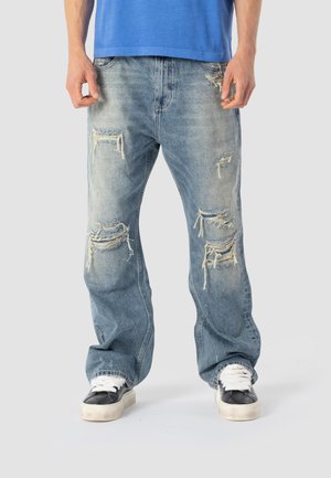 Person iført lyseblå, slidte jeans med lige ben og flossede huller, hvide og sorte sneakers samt en blå skjorte, stående på en hvid gulv.