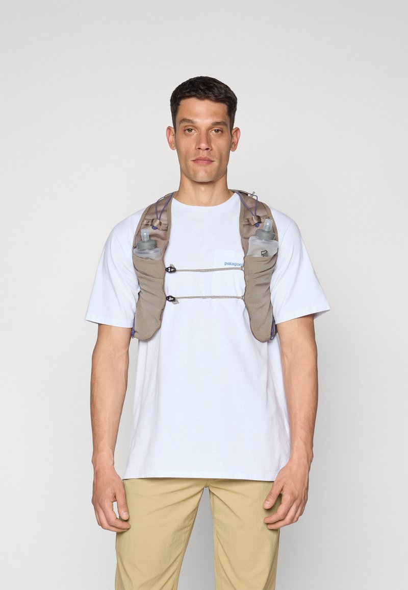 Running vest in beige fabric, featuring two water bottles, adjustable straps, and a minimal design. Worn over a white t-shirt.