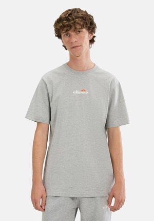 Junger Mann mit lockigem Haar, der ein hellgraues ellesse-T-Shirt und passende Shorts vor einem schlichten weißen Hintergrund trägt.