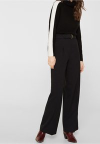 Kobieta w czarnych szerokich spodniach z wysokim stanem, czarnym golfie z białymi paskami na rękawach oraz bordowych botkach na obcasie do kostki.