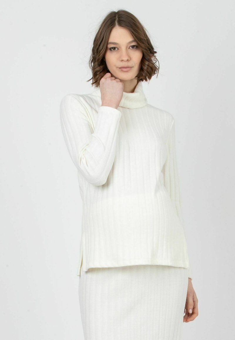 Valkoinen ribbineuleinen turtleneck-neulepusero pitkillä hihoilla, jossa on sivuaukko. Kangas näyttää pehmeältä ja sileältä.