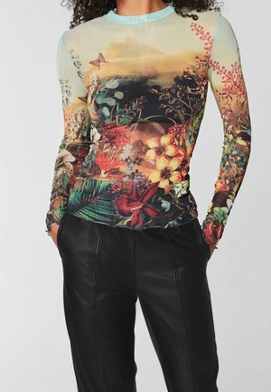Vrouw draagt een doorzichtig top met lange mouwen met een kleurrijke bloemen- en vlinderprint, gecombineerd met zwarte leren broek, handen in de zakken.